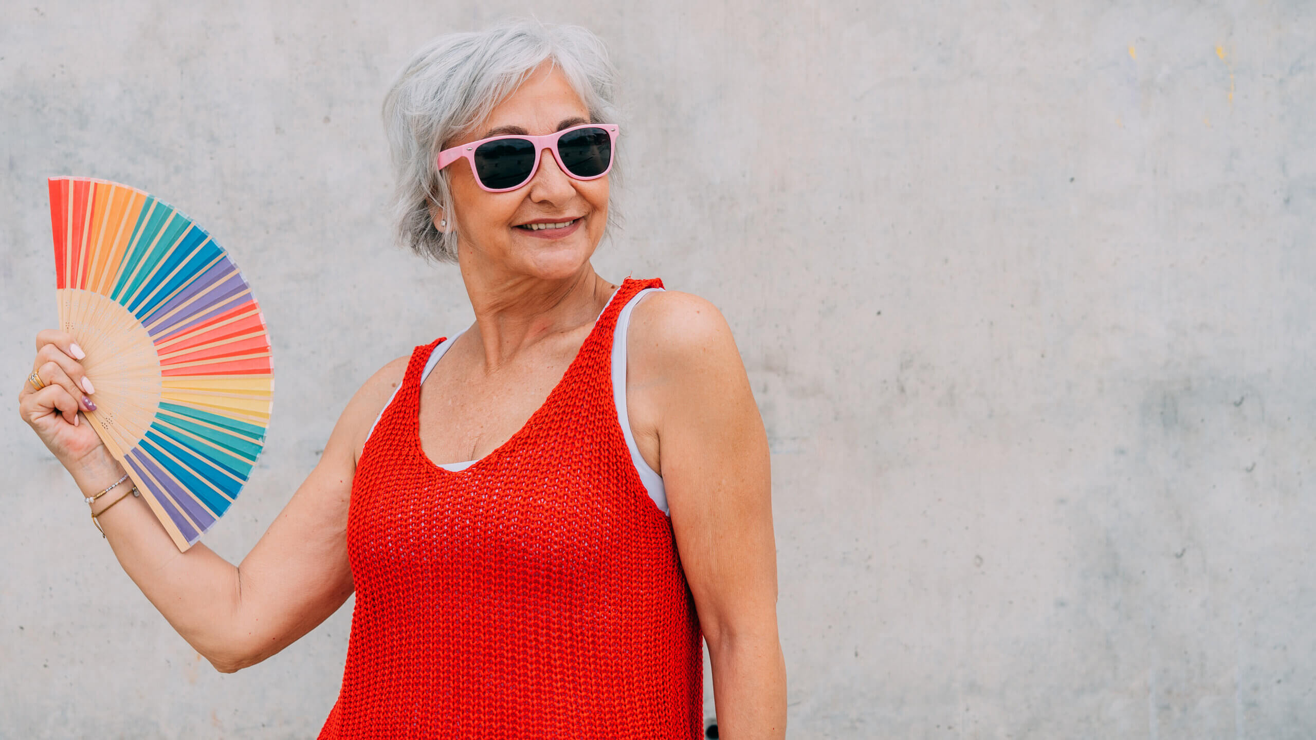 Senior female in red knitted top and pink sunglasses using multicolored striped hand fan during summer day against gray concrete wall