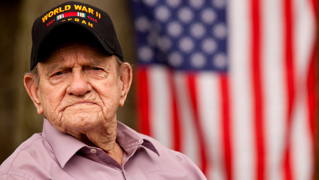 Stern male veteran in front of United States flag wearing WWII Veteran ball cap